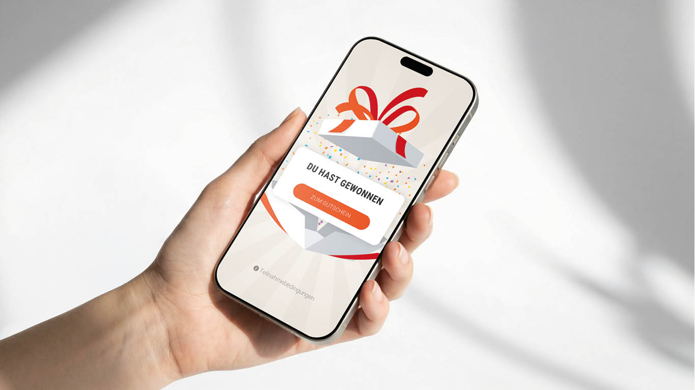 Eine Hand hält ein Smartphone, auf dessen Bildschirm eine Grafik der Masterclass Gamification-Geschenkbox zu sehen ist, über der der Text DU HAST GEWONNEN über einer Schaltfläche mit der Aufschrift ZUM GUTSCHEIN steht.